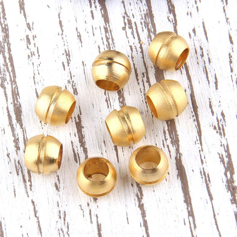 May include: Eight gold-colored metal beads with a grooved design. The beads are arranged on a white wooden surface.