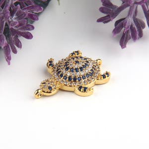 May include: Gold-tone turtle charm with blue and white crystal accents. The turtle is detailed with a textured shell and has a loop for attaching to a necklace or bracelet.
