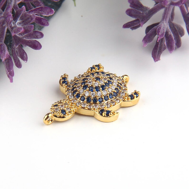 May include: Gold-tone turtle charm with blue and white crystal accents. The turtle is detailed with a textured shell and has a loop for attaching to a necklace or bracelet.