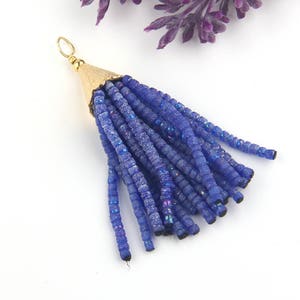 May include: A decorative tassel pendant featuring multiple strands of small, iridescent blue beads attached to a textured, gold-toned metal cone cap with a small circular jump ring at the top for hanging or attachment to jewelry pieces.