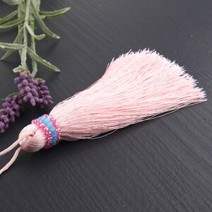 May include: A light pink silk tassel featuring a decorative band of small, faceted beads in alternating rows of bright pink and light blue. The tassel has a smooth, gathered top section and a long, flowing fringe of fine, soft threads.