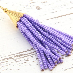 May include: A beaded tassel pendant featuring multiple strands of small, round, light purple beads. The top of the tassel is secured with a textured, gold-toned metal cap that includes a small loop for attachment at the very top of the piece.