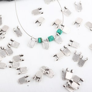 May include: Silver-toned metal jewelry findings, including rectangular pendants with small loops and turquoise beads. The pendants are scattered on a white surface, some strung on a thin wire.