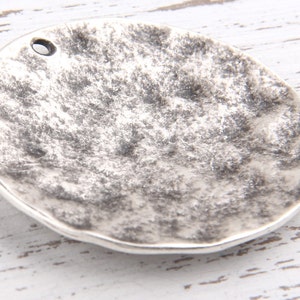 Puede incluir: Un dije redondo de metal plateado con una superficie texturizada y un pequeño agujero en la parte superior.