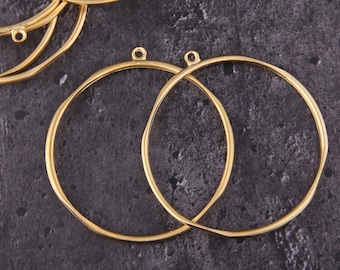 Puede incluir: Dos aros de metal de color dorado con un pequeño bucle en la parte superior de cada aro. Los aros están sobre una superficie texturizada gris oscuro.