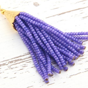 May include: A purple beaded tassel pendant featuring numerous strands of small, uniform purple glass beads attached to a textured, conical gold-tone metal cap with a small circular jump ring at the top for attachment to jewellery or accessories.