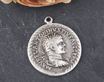 May include: A silver-colored round pendant with a detailed relief of a man's profile and Latin text around the edge. The pendant has a loop at the top for hanging and is likely a historical or antique-inspired piece.