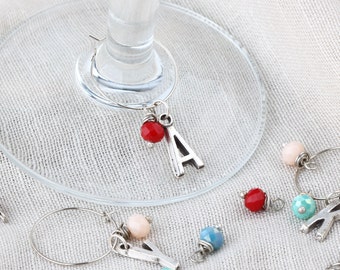 May include: Silver wine glass charms with colorful beads and letter charms. The charms are hanging from a wine glass.