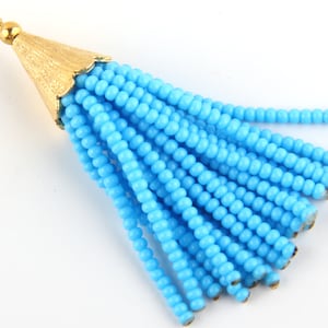 May include: A bright blue beaded tassel pendant featuring a textured gold-toned metal cap at the top. The tassel consists of multiple strands of small, uniform, round blue beads hanging from the conical cap, which includes a small circular jump ring.