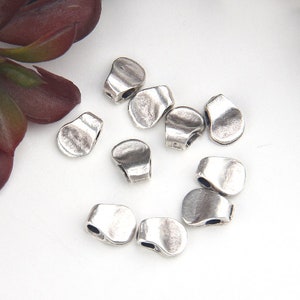 May include: A collection of silver-colored, irregular-shaped beads. Each bead has a slightly flattened, rounded form with a hole on each end. The beads are scattered on a white surface, with a hint of burgundy foliage in the corner.