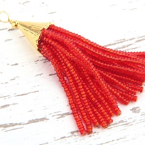 May include: A red beaded tassel pendant featuring a gold-toned metal cap with a small circular jump ring at the top. The tassel is composed of numerous thin strands of small, translucent red glass beads hanging from the metallic cone-shaped cap.
