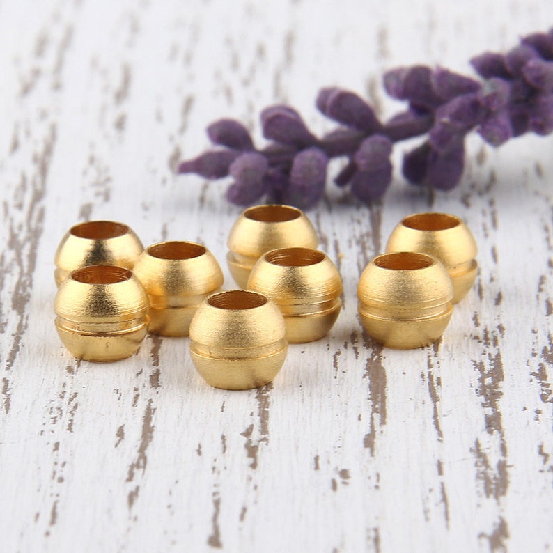 May include: Eight gold-colored metal beads, each with a slightly rounded, cylindrical shape. The beads are arranged in a semi-circle on a white wooden surface.