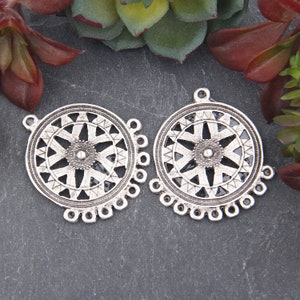 Può includere: Due ciondoli circolari color argento con intricati disegni geometrici. Ogni ciondolo presenta un motivo centrale a stella, circondato da una serie di triangoli e un bordo smerlato con piccoli anelli. Ideale per la creazione di gioielli.