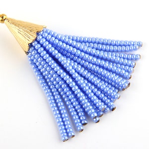 May include: A light blue beaded tassel pendant featuring a gold-toned metal cap with a textured, fluted design. The tassel consists of multiple strands of small, uniform, glossy light blue beads attached to a small gold-toned circular jump ring.
