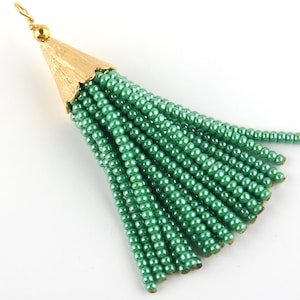 May include: A decorative tassel pendant featuring a textured gold-tone metal cap with a small loop at the top. Attached to the cap are numerous strands of small, shiny, metallic green seed beads that hang down in a fan-like arrangement.