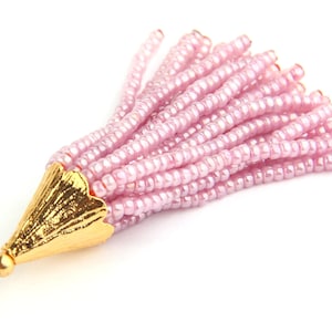 May include: A tassel pendant featuring numerous strands of light pink, iridescent seed beads attached to a textured, gold-toned metal cone cap. The cap is topped with a small, round gold bead and a circular wire loop for attachment purposes.