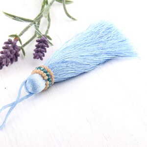 May include: A light blue silk tassel featuring a decorative band of small, round, beige beads and shimmering teal crystal beads. The tassel includes a matching light blue cord loop at the top for hanging, creating a soft and textured appearance.