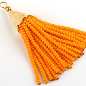 Pode incluir: Uma borla decorativa com numerosos fios de missangas cor de laranja brilhante fixados a uma tampa cónica de metal dourado texturado. A tampa inclui um pequeno laço circular no topo para pendurar, proporcionando um ponto de fixação seguro.