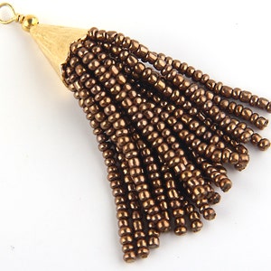 May include: A tassel pendant featuring multiple strands of metallic bronze seed beads attached to a textured gold-tone metal cone cap. The cap includes a small circular loop at the top for attachment, creating a dangling jewellery component.