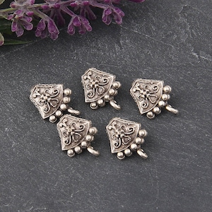 May include: Five silver-toned metal shield-shaped charms with ornate floral designs and small ball accents. Each charm has a small loop at the bottom. The charms are arranged on a dark gray surface, with purple flowers in the background.