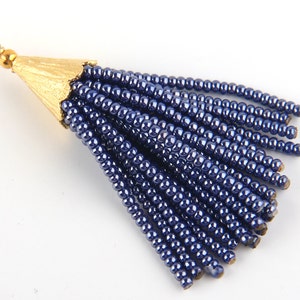 May include: A decorative tassel pendant featuring multiple strands of metallic dark blue seed beads attached to a textured gold-toned metal cap with a small circular jump ring at the top for hanging or attachment to jewellery or accessories.