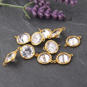 May include: A group of ten gold-toned round pendant charms with clear crystal stones. The charms have a loop on the top for attaching to a necklace or bracelet.