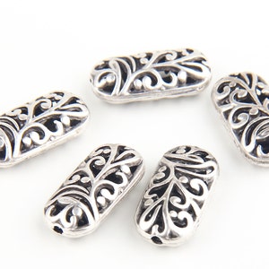 May include: Five silver-colored, oval-shaped beads with intricate, openwork floral designs. The beads are arranged on a white background, showcasing their detailed patterns and metallic sheen. These decorative beads are suitable for jewelry making.