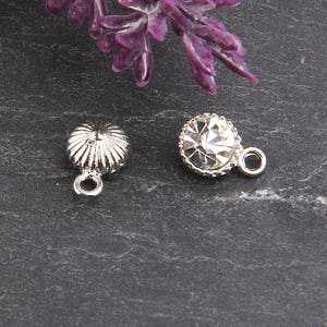 May include: Two silver-toned charms. One charm is a small, ridged, flower-like shape with a loop. The other is a round charm with a clear, faceted crystal center and a silver-toned, beaded edge, also with a loop.