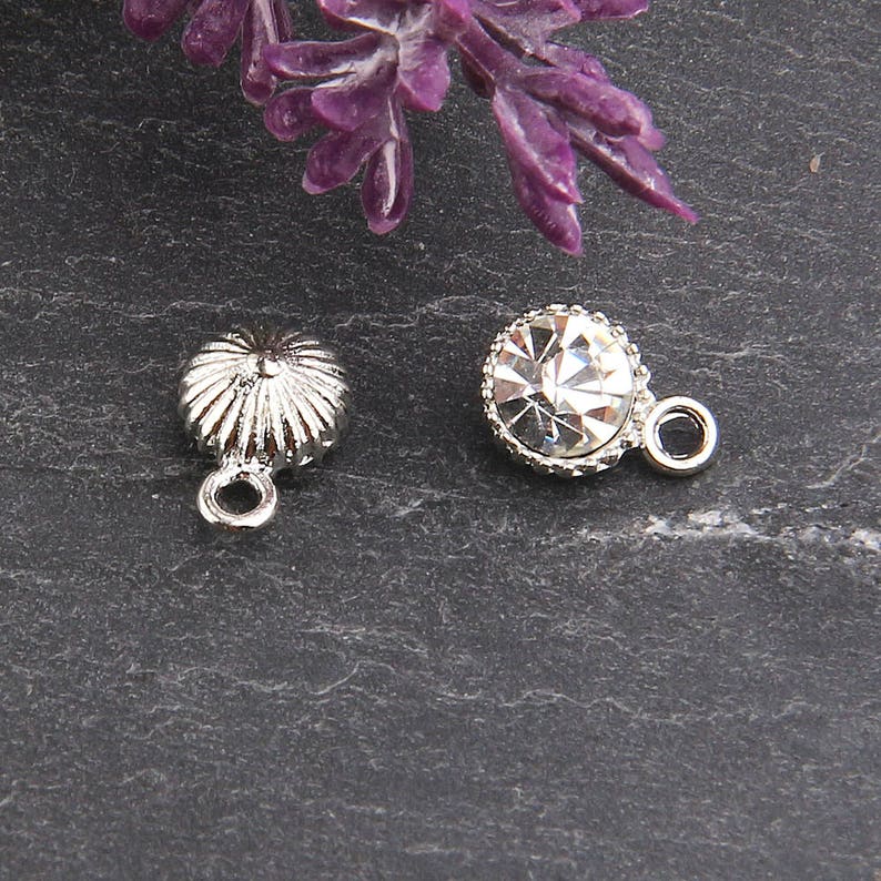 May include: Two silver-toned charms. One charm is a small, ridged, flower-like shape with a loop. The other is a round charm with a clear, faceted crystal center and a silver-toned, beaded edge, also with a loop.