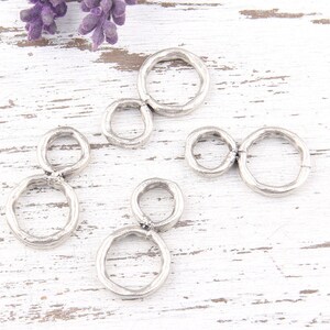 Puede incluir: Encantos de metal plateado con doble anillo. Cada dije presenta dos anillos conectados de forma irregular. Los dijes están esparcidos sobre una superficie de madera blanca desgastada, con una ramita de flores moradas en el fondo.