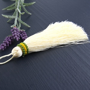May include: A cream-coloured silk tassel featuring a decorative band of small green and yellow beads at the top. The tassel includes a matching cream-coloured cord loop for hanging, with a smooth, rounded knot securing the fine, flowing threads.