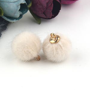 May include: Two round, cream-colored faux fur pom-pom charms. Each fluffy sphere features a small, shiny gold-toned metal loop at the top, designed for attachment to jewelry, accessories, or craft projects. The texture is soft and dense.