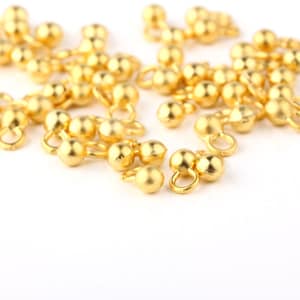 May include: Close-up of numerous small, gold-colored jewelry findings. Each finding features a rounded bead shape with a small loop attached. The findings are scattered across a white surface, creating a bright and reflective appearance.