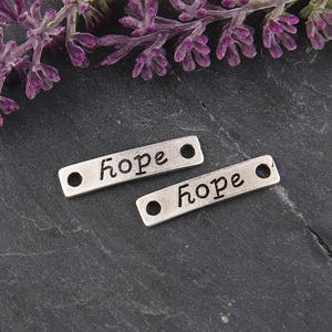 Könnte beinhalten: Zwei silberne, rechteckige Anhänger mit dem Wort "hope" in Schreibschrift. Jeder Anhänger hat an jedem Ende ein Loch. Die Anhänger liegen auf einer dunkelgrauen Oberfläche, mit lila Blumen im Hintergrund.