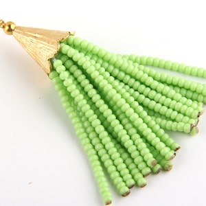 May include: A light green beaded tassel pendant featuring multiple strands of small, uniform round beads attached to a textured, gold-toned metal cap with a small circular loop at the top for hanging or attachment to jewellery components.
