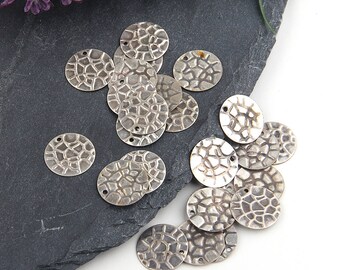 Peut inclure: Un ensemble de breloques rondes en métal argenté texturé, avec un motif en mosaïque. Chaque breloque comporte deux petits trous pour l'enfilage. Les breloques sont disposées sur une surface d'ardoise gris foncé, avec des fleurs violettes en arrière-plan.