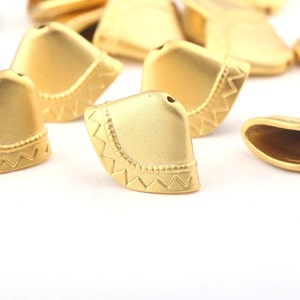 May include: A collection of gold-colored metal beads. Each bead has a hat-like shape with a decorative zigzag pattern along the edge. The beads are scattered on a white surface, with some in focus and others blurred.