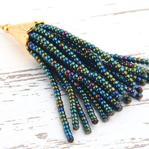 May include: A decorative tassel pendant featuring multiple strands of iridescent dark blue and green seed beads attached to a textured gold-tone metal cone cap with a small loop at the top for hanging or attachment to jewellery pieces.