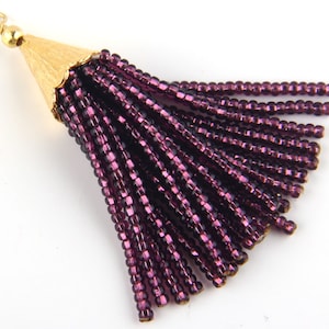 May include: A decorative tassel pendant featuring a gold-toned metal cone cap with a small circular jump ring at the top. The tassel consists of multiple strands of deep purple, reflective glass seed beads hanging from the metal cap.