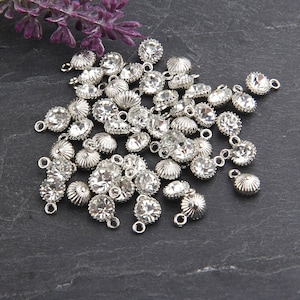 May include: A collection of silver-toned rhinestone charms with clear, faceted crystals. The charms have a decorative, scalloped edge and small loops for attaching to jewelry or other crafts. A sprig of purple flowers is in the upper left corner.