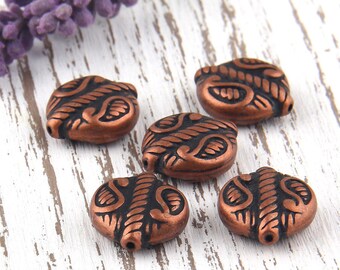 May include: Five copper-colored, oval-shaped beads with a dark, etched design. The beads have a hole on each end for stringing. A sprig of purple lavender is in the background, resting on a white, distressed wood surface.