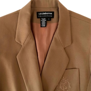 K&ouml;nnte beinhalten: Ein beigefarbener Blazer mit steigendem Revers und pfirsichfarbenem Innenfutter. Das Innenetikett zeigt Liz Claiborne Collection, Gr&ouml;&szlig;e 6. Ein gesticktes Wappen mit den Buchstaben LC ist auf der unteren Vordertasche zu sehen.
