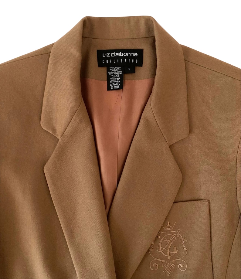 K&ouml;nnte beinhalten: Ein beigefarbener Blazer mit steigendem Revers und pfirsichfarbenem Innenfutter. Das Innenetikett zeigt Liz Claiborne Collection, Gr&ouml;&szlig;e 6. Ein gesticktes Wappen mit den Buchstaben LC ist auf der unteren Vordertasche zu sehen.