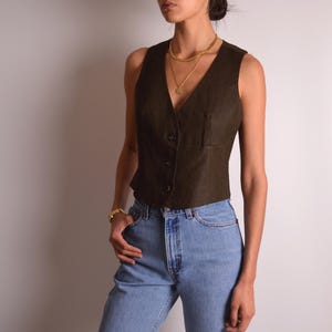 Könnte beinhalten: Eine dunkelbraune Weste mit V-Ausschnitt und Knopfverschluss. Die Weste hat eine kleine Tasche auf der linken Seite und wird mit hellblauen Jeans getragen. Eine goldene Halskette und ein Armband sind ebenfalls sichtbar.