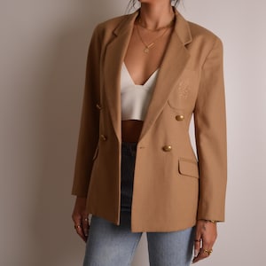 May include: A camel-colored double-breasted blazer featuring gold-toned buttons, a notched lapel, and a subtle embroidered crest detail on the left chest pocket area. The blazer is worn over a white cropped top and light blue denim jeans.