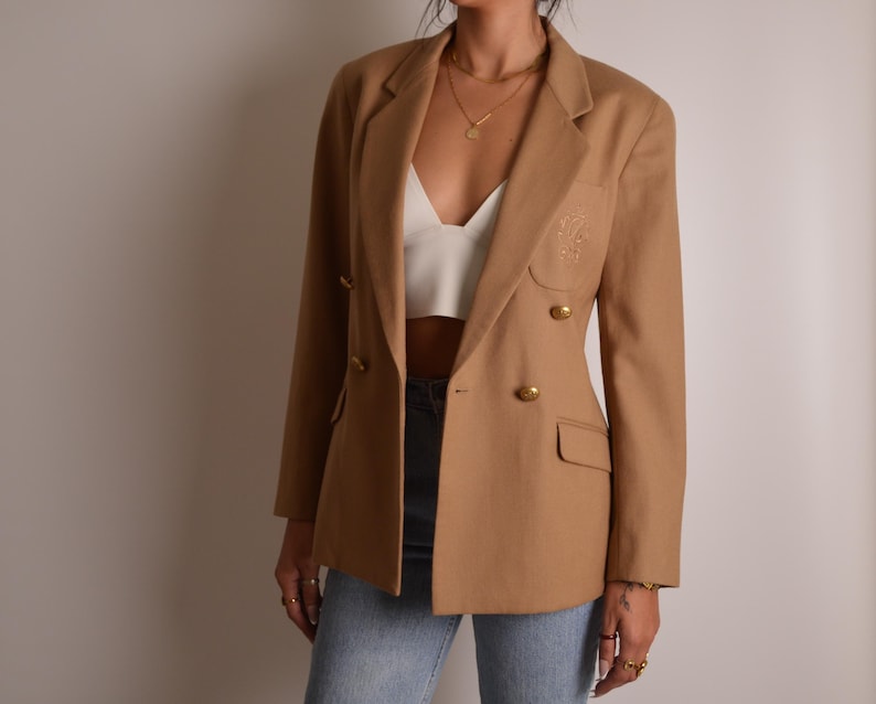 K&ouml;nnte beinhalten: Ein kamelfarbener zweireihiger Blazer mit goldfarbenen Kn&ouml;pfen, steigendem Revers und einem dezenten gestickten Wappen-Detail im Bereich der linken Brusttasche. Der Blazer wird &uuml;ber einem wei&szlig;en bauchfreien Top und einer hellblauen Jeans getragen.