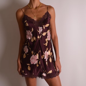May include: Burgundy satin slip dress with a floral pattern in pink and cream. The dress features spaghetti straps, a lace trim neckline, and a matching lace hem. A delicate, feminine garment.