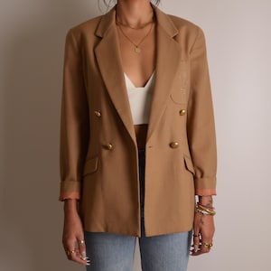 K&ouml;nnte beinhalten: Ein beigefarbener zweireihiger Blazer mit goldfarbenen Kn&ouml;pfen, fallendem Revers und zwei Pattentaschen auf der Vorderseite. Die &Auml;rmel sind umgeschlagen und zeigen ein hellrosa Innenfutter, zudem ist ein dezentes Stickemblem sichtbar.