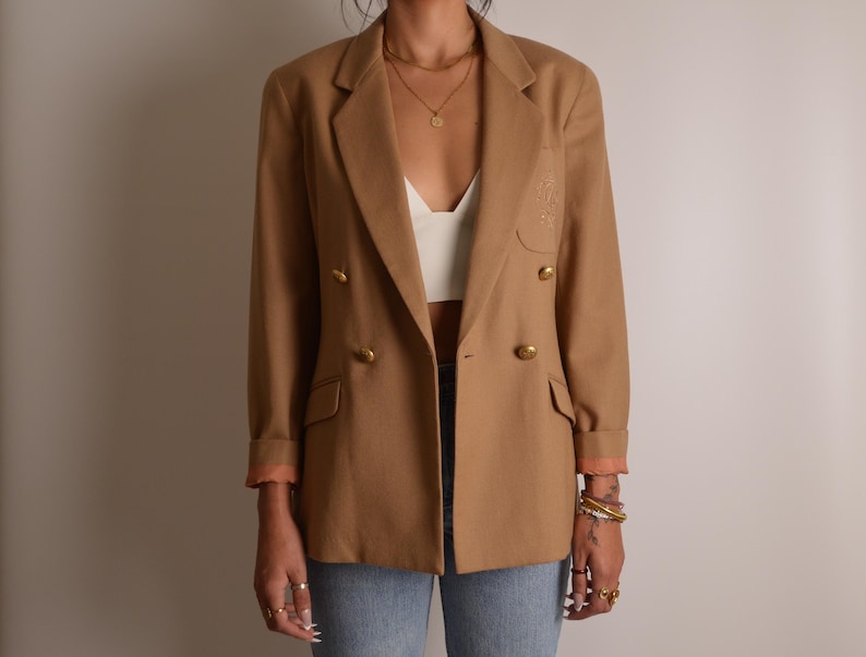 K&ouml;nnte beinhalten: Ein beigefarbener zweireihiger Blazer mit goldfarbenen Kn&ouml;pfen, fallendem Revers und zwei Pattentaschen auf der Vorderseite. Die &Auml;rmel sind umgeschlagen und zeigen ein hellrosa Innenfutter, zudem ist ein dezentes Stickemblem sichtbar.
