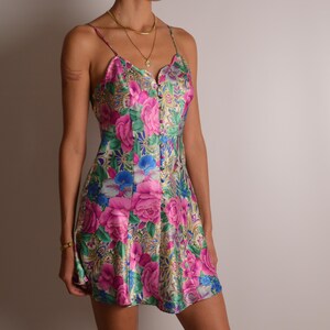 May include: A short, sleeveless dress featuring a vibrant floral pattern in shades of bright pink, blue, and green on a light background. The dress has thin spaghetti straps, a V-neckline, and a vertical row of small buttons down the center front.